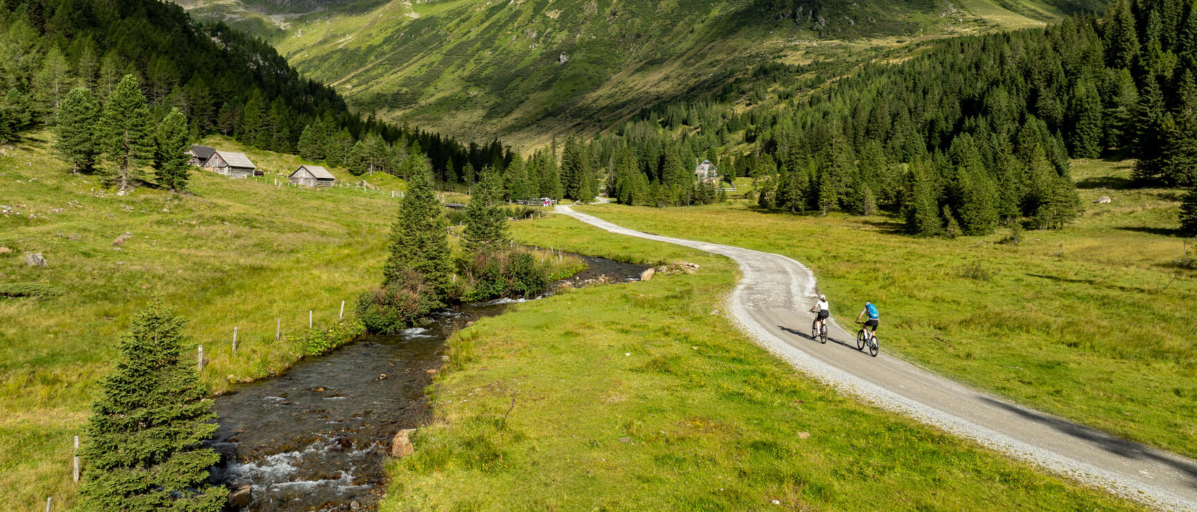 Two cyclists on a gravel path in an alpine valley with a stream and wooden huts on the left, green mountains in the background.