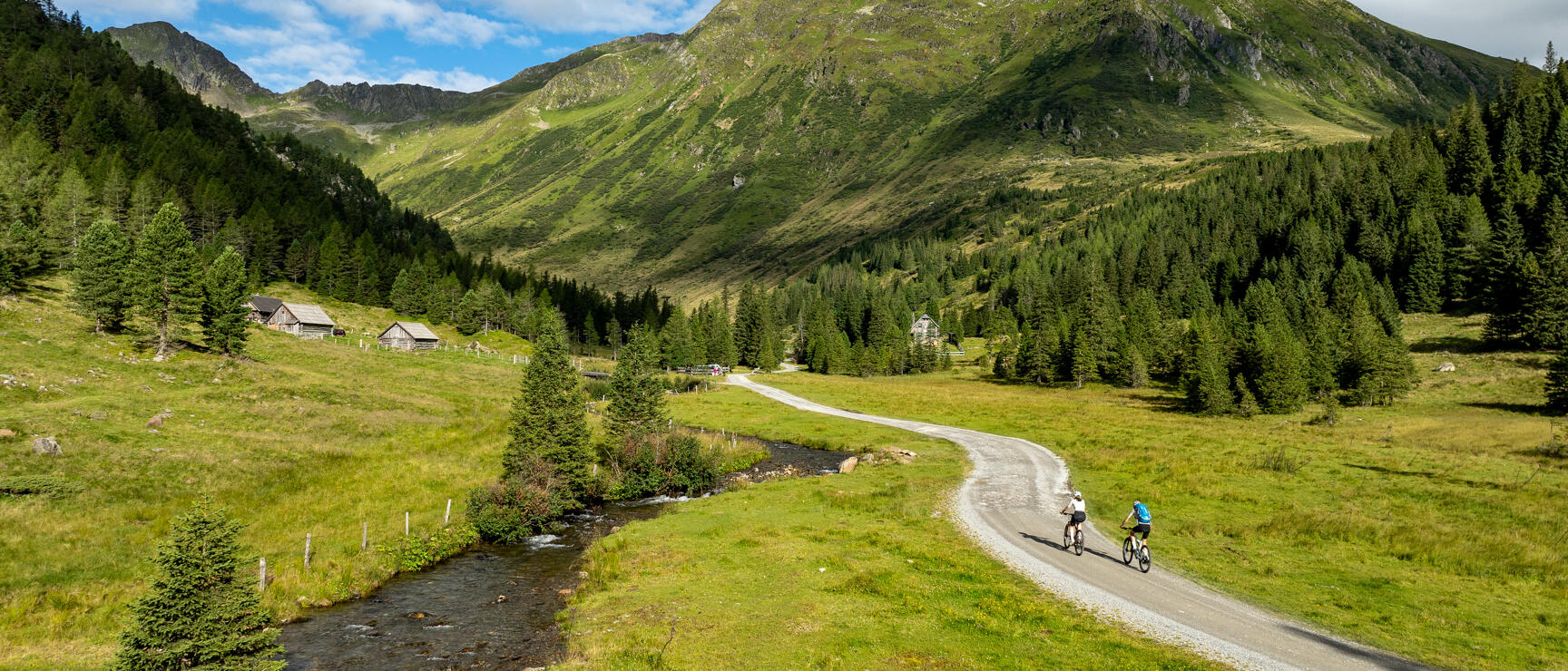 Zwei Radfahrer auf Schotterweg im Almtal, Bach und Holzhütten links, grüne Berge im Hintergrund.