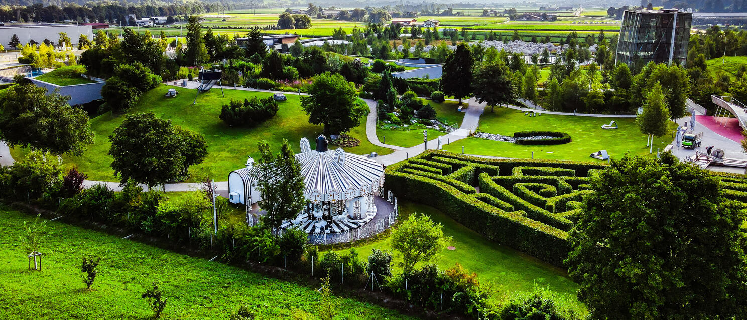 Aerial view of Swarovski Crystal Worlds in Wattens: hedge maze, pavilion, glass tower, mountain panorama in background.