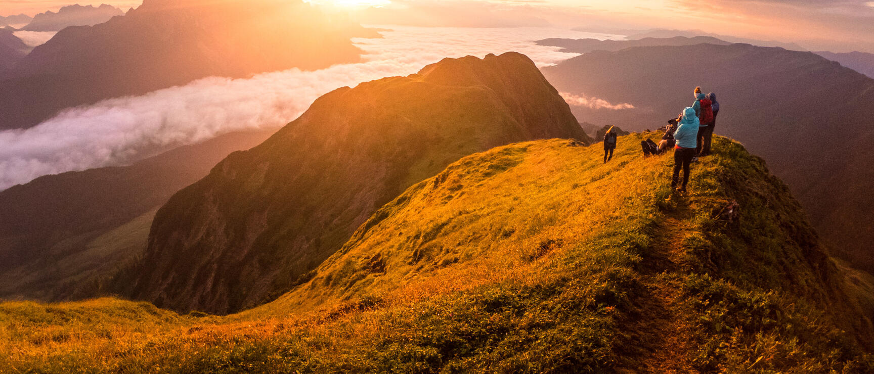 Multiple people on a mountain ridge at sunset, sea of clouds and mountain panorama in the background.