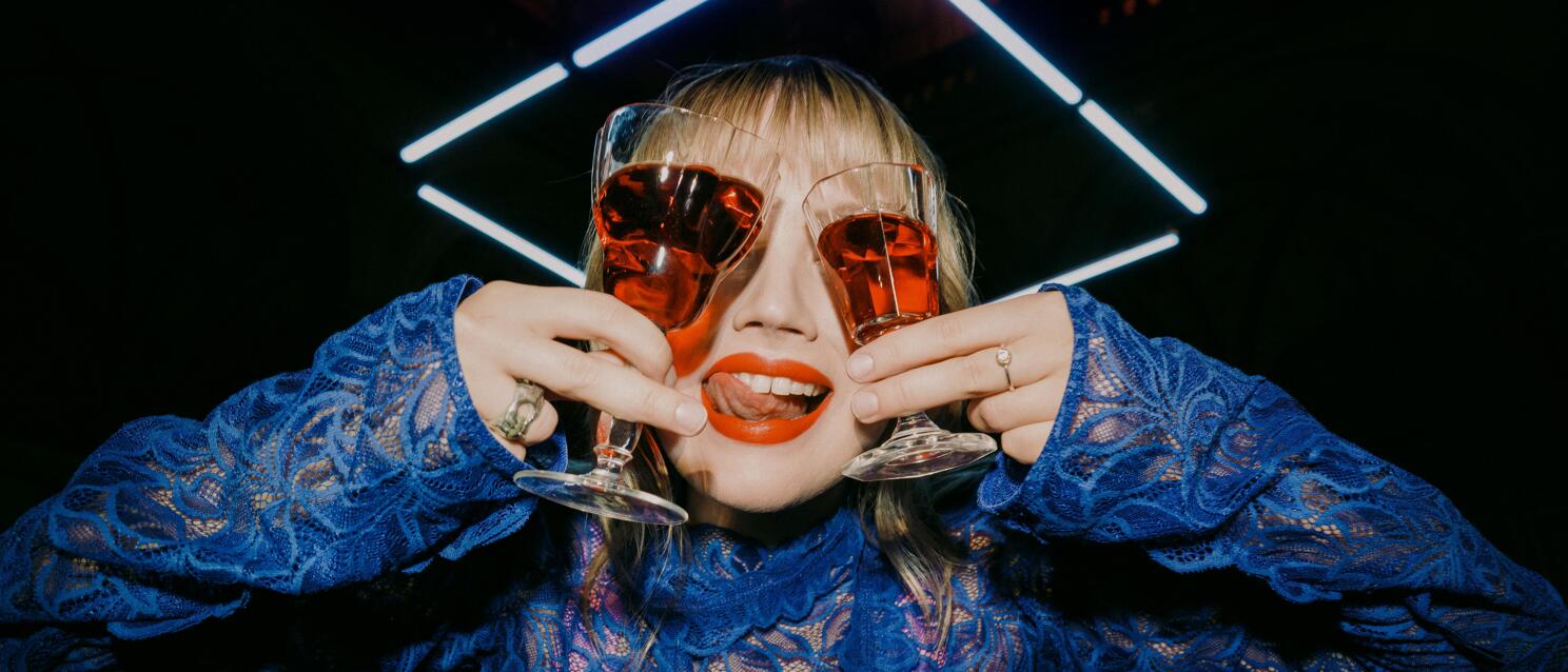 Woman in blue lace blouse holding two red wine glasses in front of her eyes, neon light and vault in background.