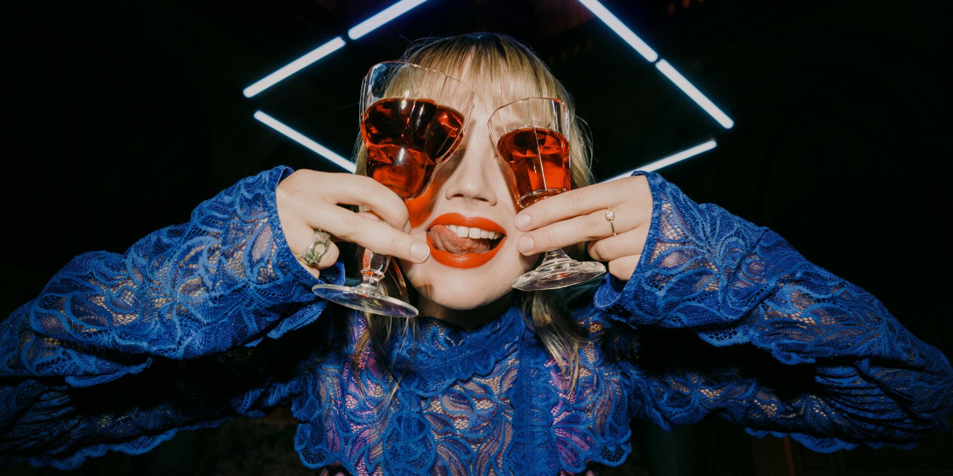 Woman in blue lace blouse holding two red wine glasses in front of her eyes, neon light and vault in background.