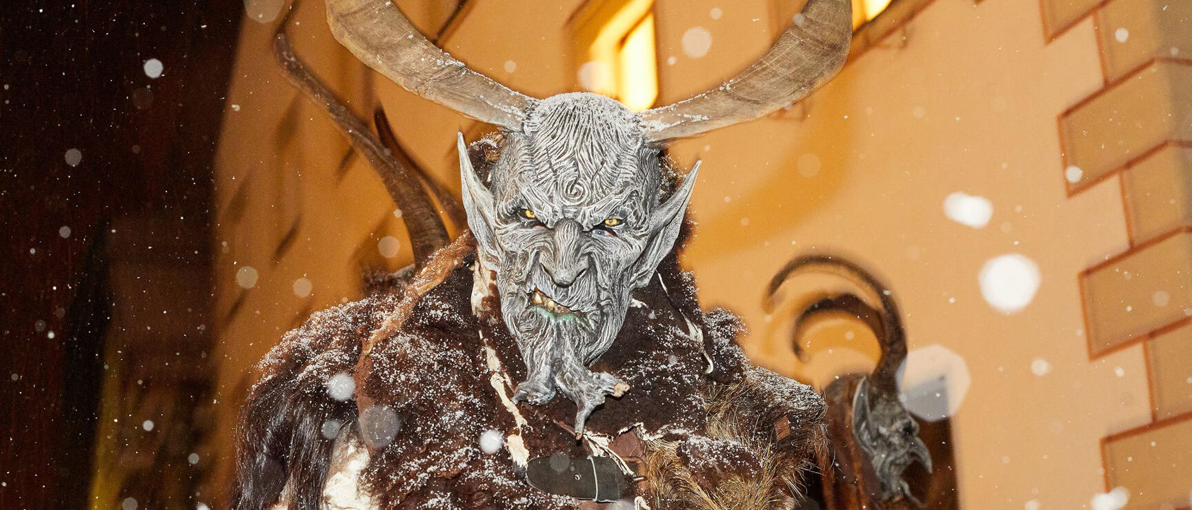 Krampus with grey mask, horns and fur costume in snowfall, race number 372, building in background.