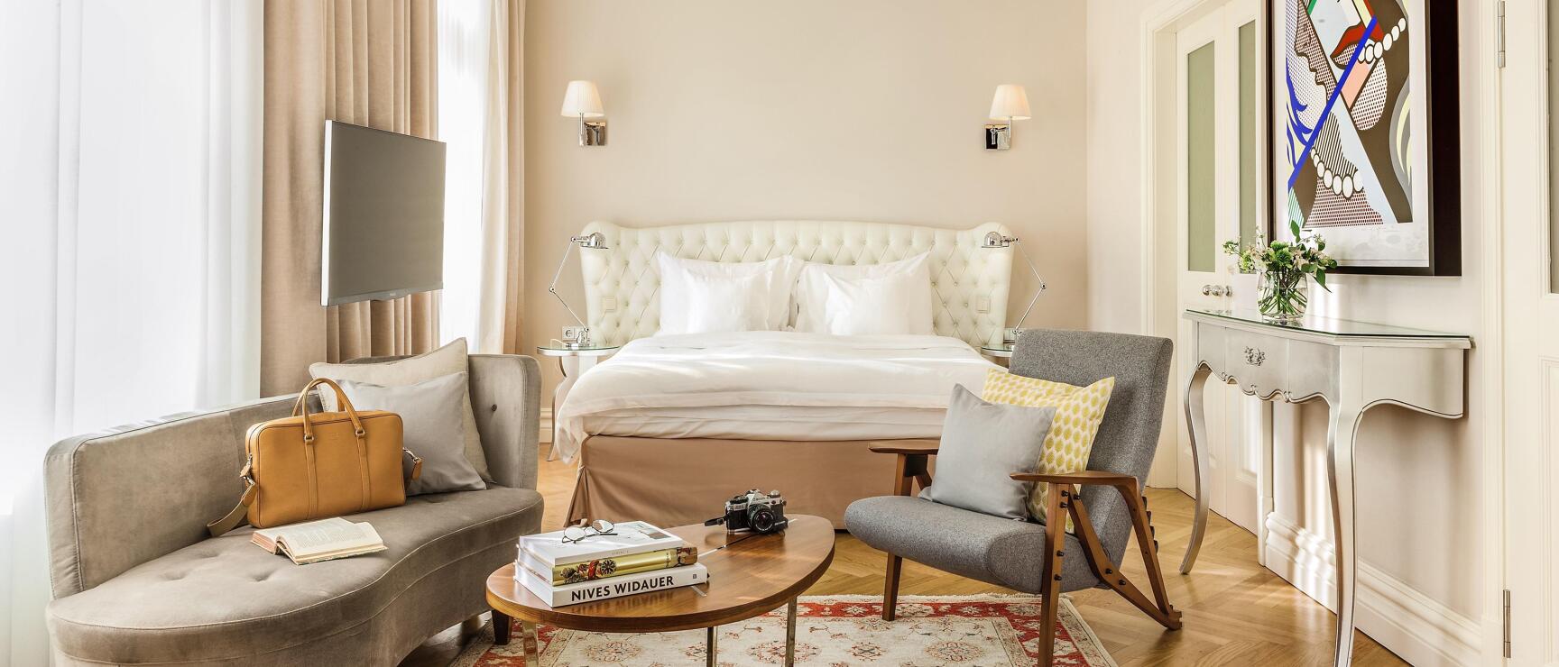 Elegant hotel room with white upholstered bed, seating area, wooden table, oriental rug, and colourful artwork on the wall.