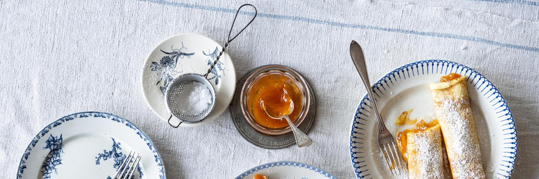 Pancakes with powdered sugar on blue-and-white plates, jam jar and powdered sugar sifter beside them.