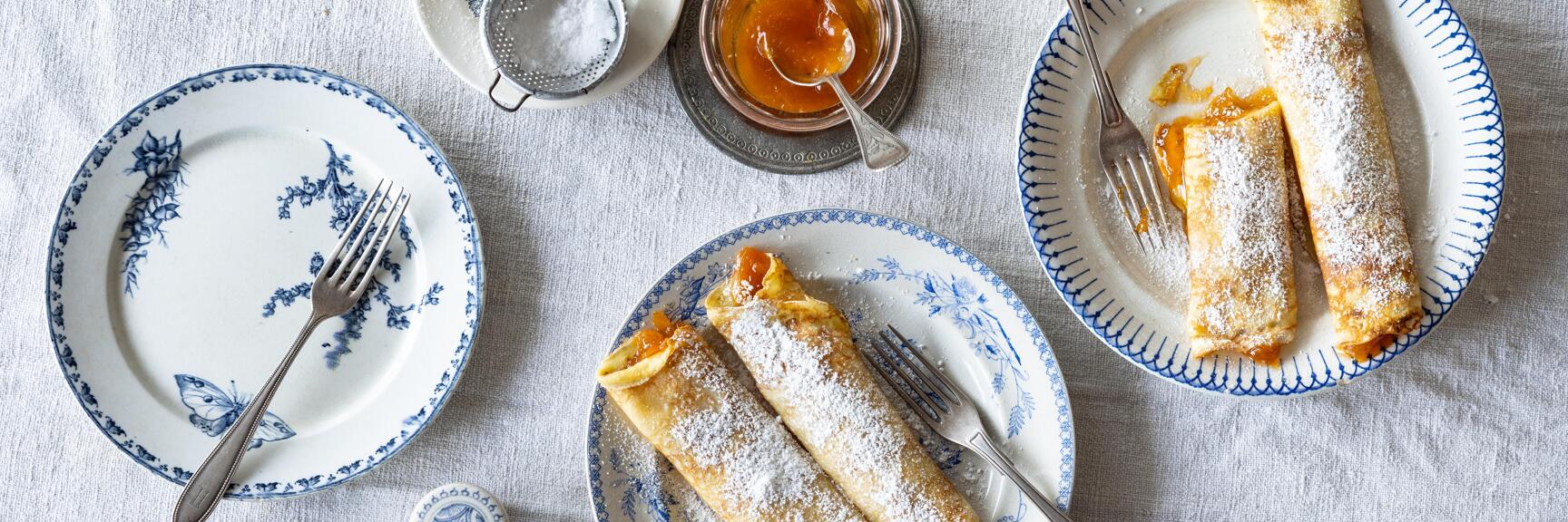 Pancakes with powdered sugar on blue-and-white plates, jam jar and powdered sugar sifter beside them.