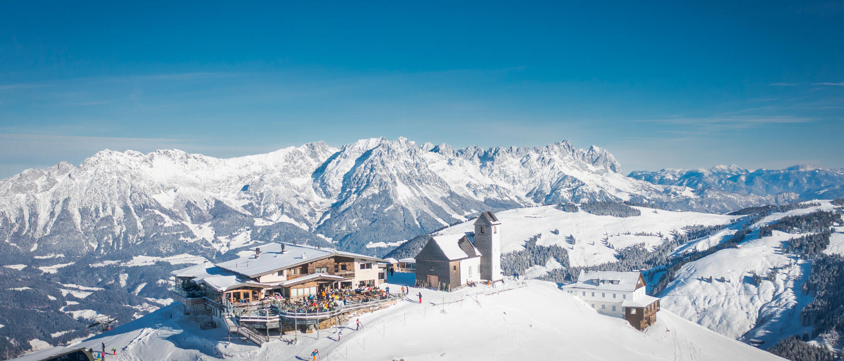 Snowy mountain peak with ski lodge, small chapel and panoramic view of snow-covered Alpine range.
