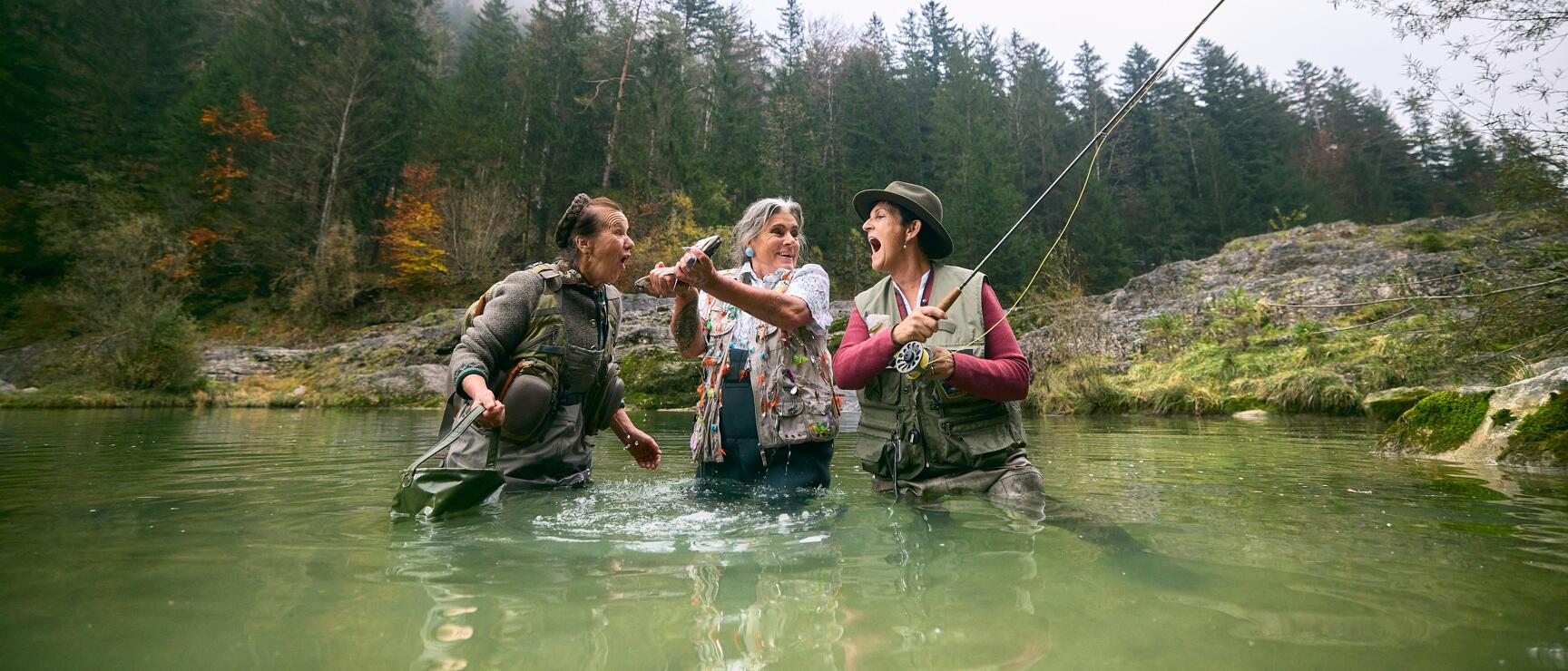 Three women with fishing rods celebrating in a river, coniferous forest and rocks in background.