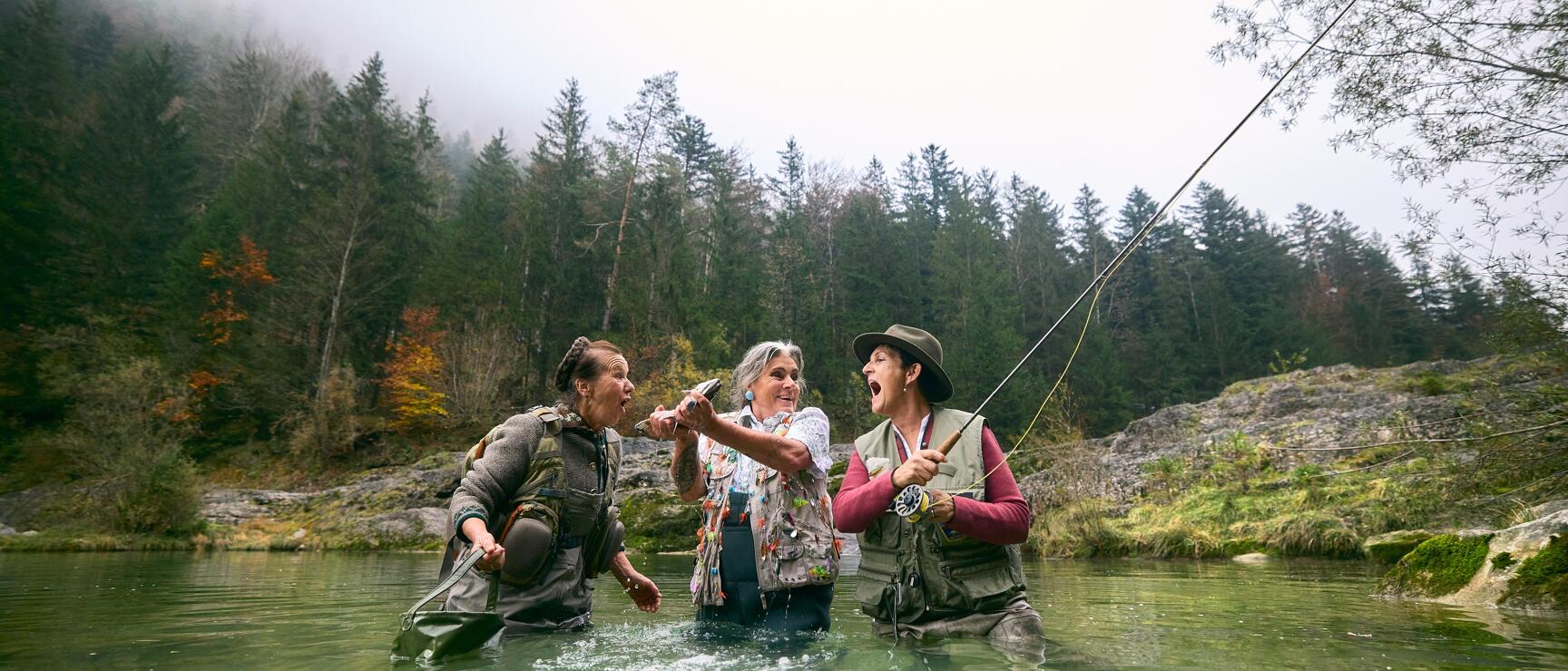 Drei Frauen mit Angelruten stehen jubelnd in einem Fluss, Nadelwald und Felsen im Hintergrund.