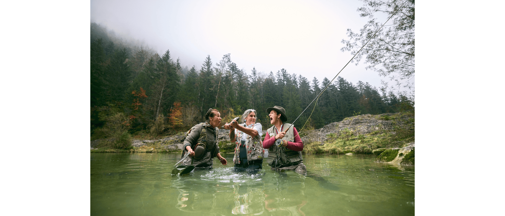 Drei Frauen mit Angelruten stehen jubelnd in einem Fluss, Nadelwald und Felsen im Hintergrund.