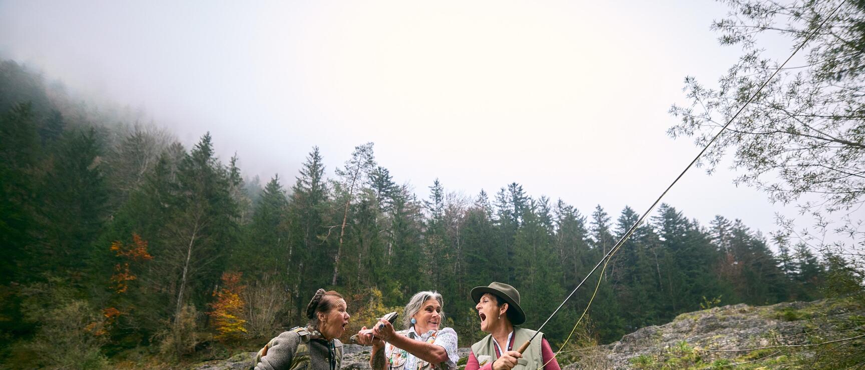 Three women with fishing rods celebrating in a river, coniferous forest and rocks in background.
