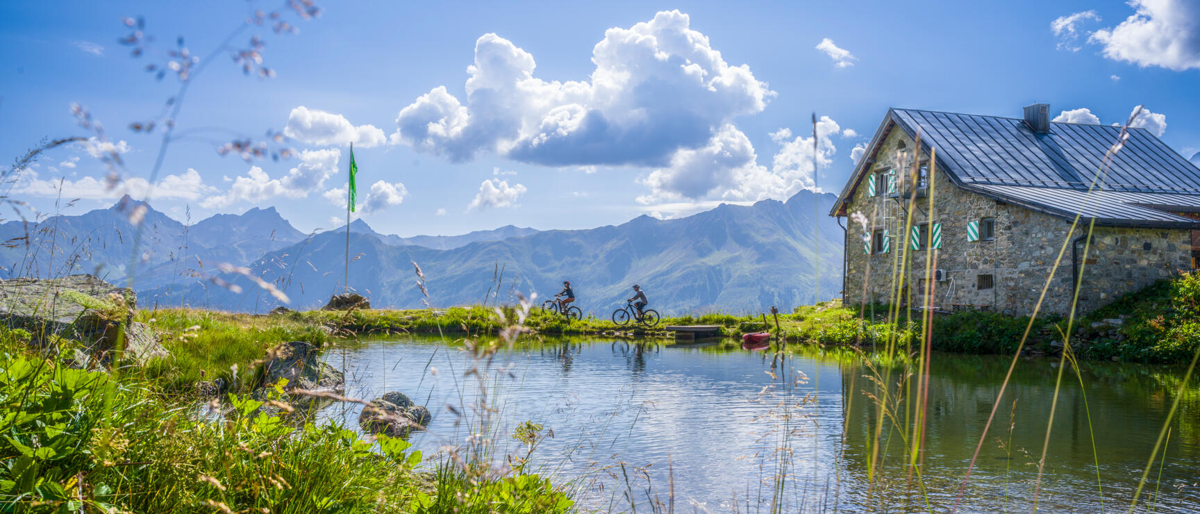 Zwei Radfahrer an einem Bergsee neben einer Steinschutzhütte, Alpenpanorama im Hintergrund.