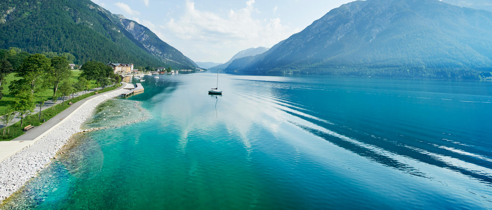 View of Lake Achensee with sailboat, shoreside path on left, village and mountains in background, Tyrol.