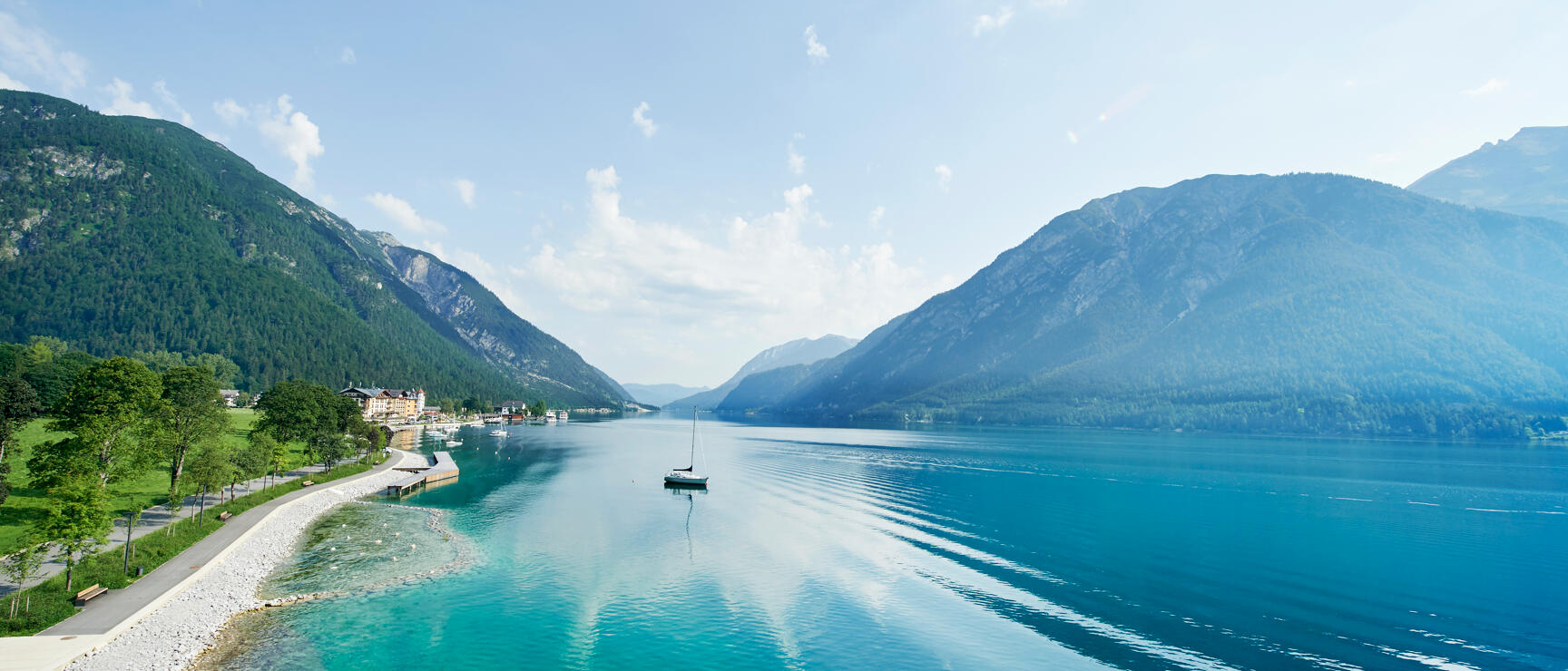 Blick auf den Achensee mit einem Segelboot, Uferweg links, Ortschaft und Bergen im Hintergrund, Tirol.
