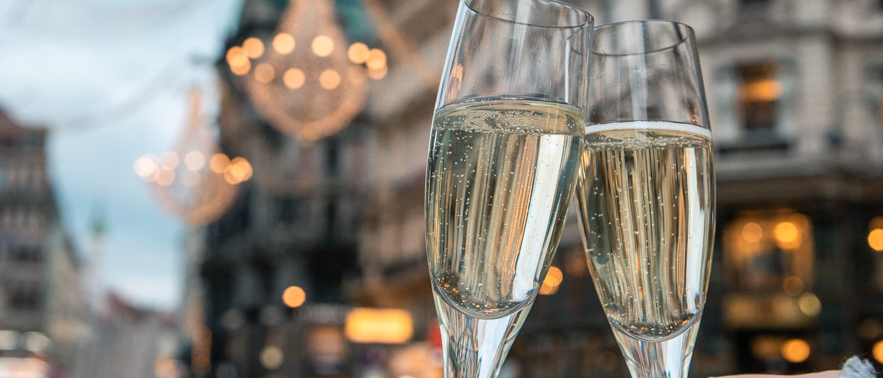Two champagne glasses clinking together, illuminated street in the background with chandeliers in Vienna city centre.