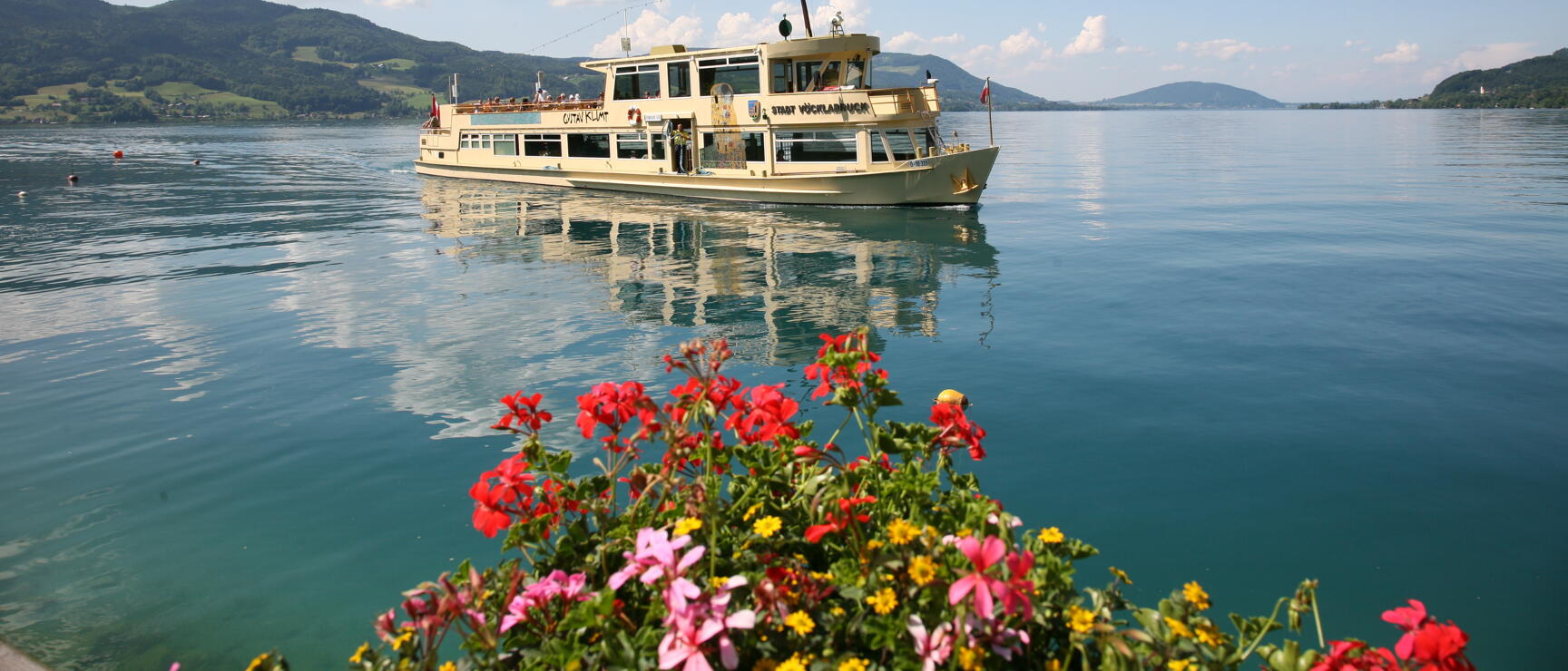 White passenger ship 'Otto Klimt' on Lake Atter, red flowers in foreground, green hills in background.