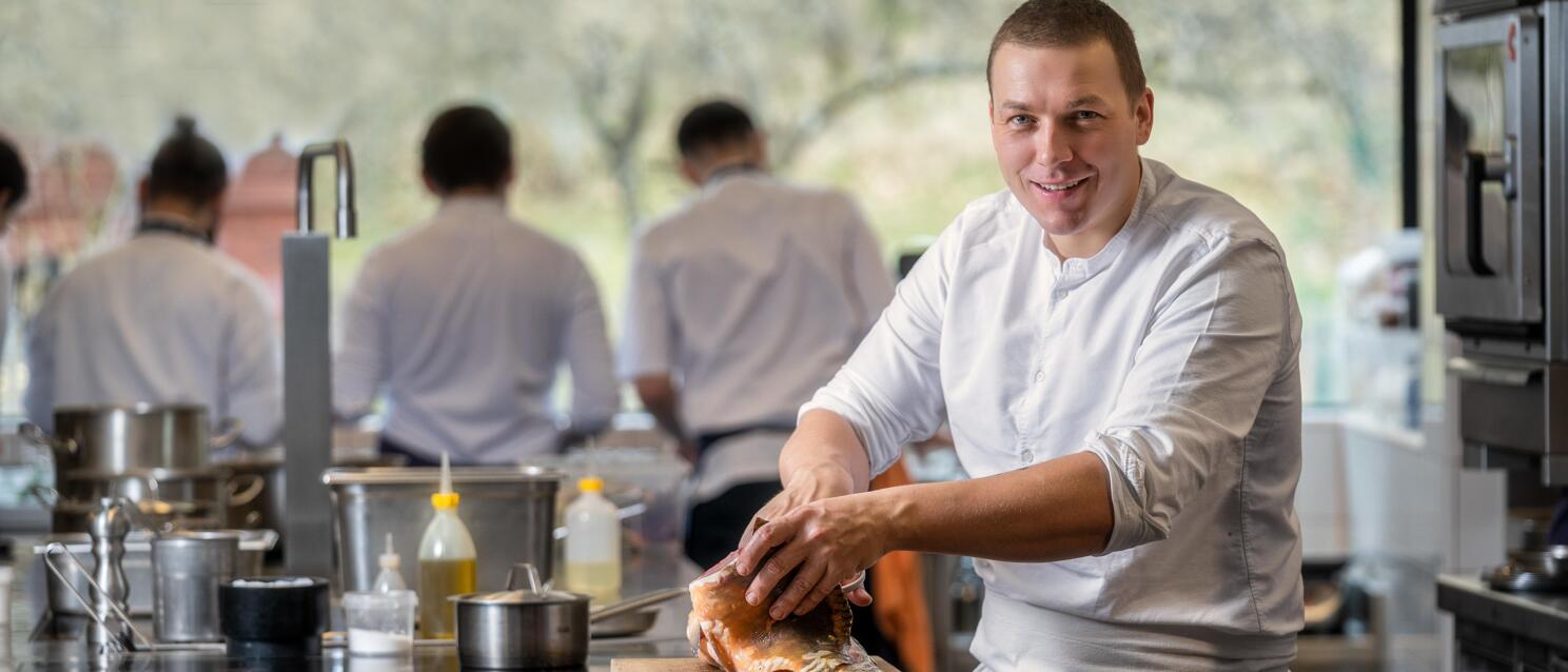 Chef in white jacket preparing fish on wooden board in professional kitchen, other chefs visible in background.