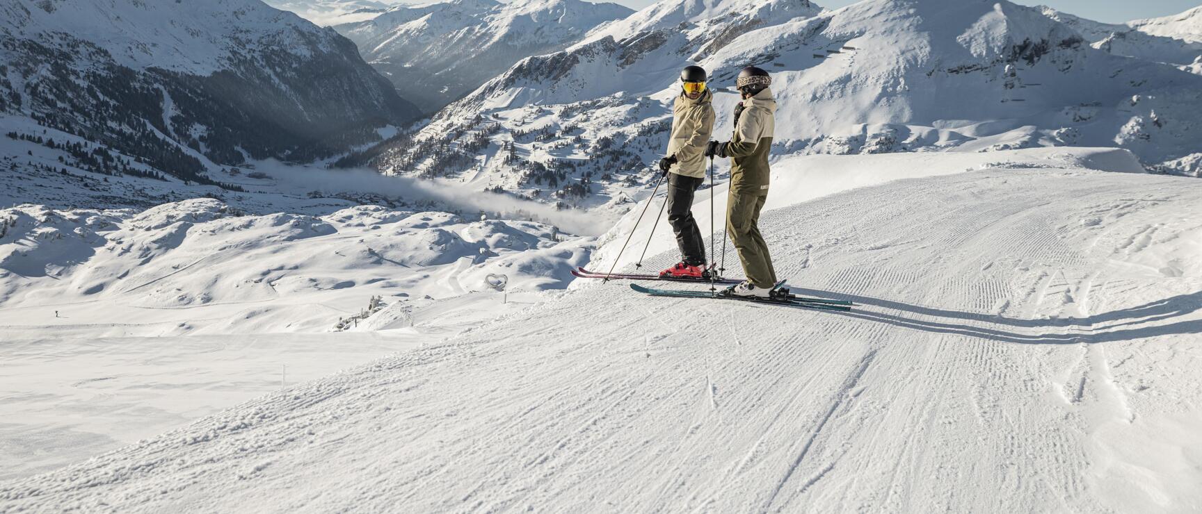 Two skiers on groomed slope, snow-covered mountain range and valley in background.