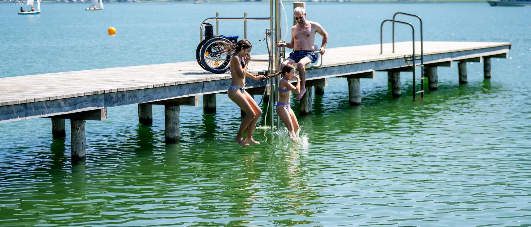 Man in wheelchair on bathing platform with two children jumping into green lake water, forested mountains in background.