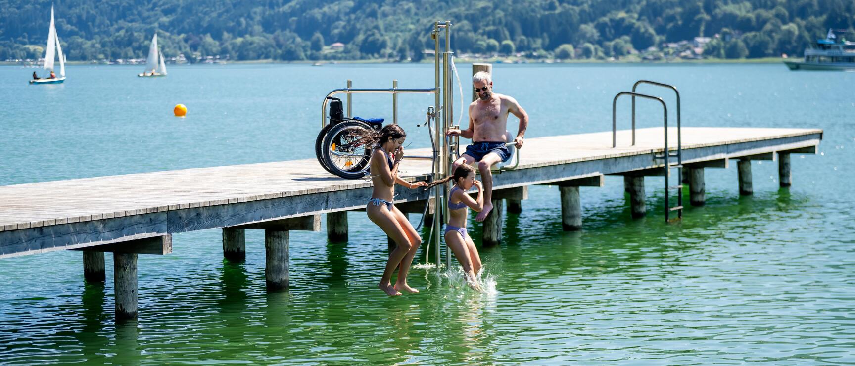 Mann auf Badesteg mit Rollstuhl, zwei Kinder springen ins grüne Seewasser, bewaldete Berge im Hintergrund.