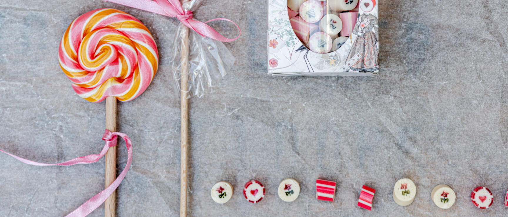 Zwei rosa-orange Lollipops, eine dekorative Bonbonschachtel und kleine runde Zuckerbonbons auf grauem Untergrund.