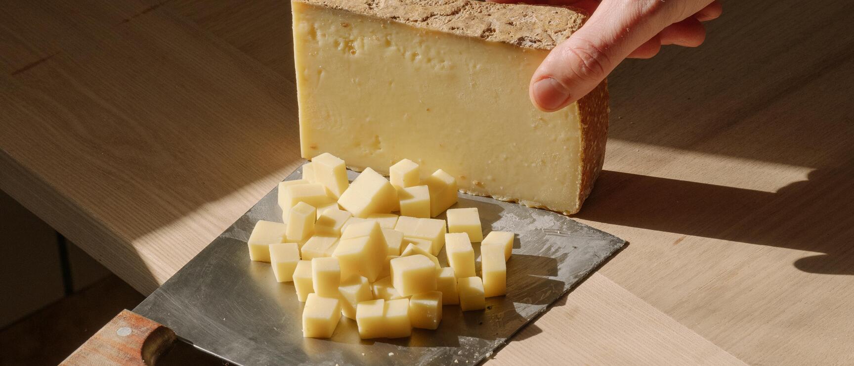 Hand holding a piece of hard cheese over a cheese slicer with diced cheese on wooden table.