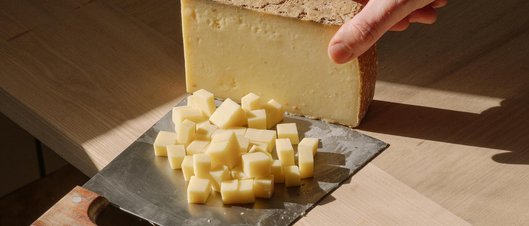 Hand holding a piece of hard cheese over a cheese slicer with diced cheese on wooden table.
