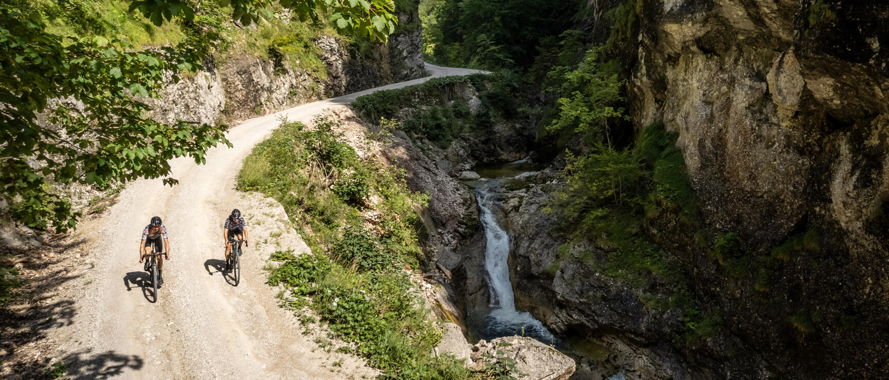 Two cyclists on gravel path through rocky canyon with waterfall, forest and blue sky.
