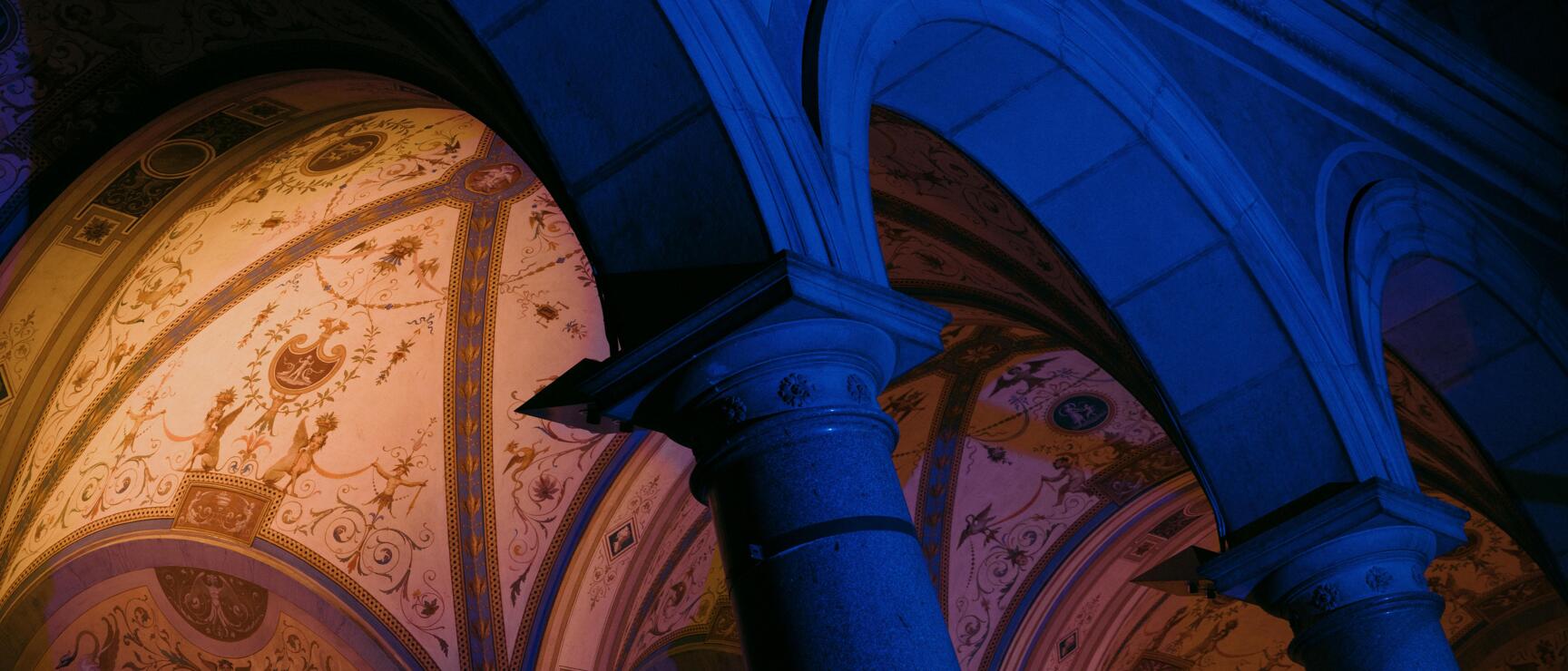 View upward into a barrel vault with frescoes, illuminated in blue and orange, columns and arches visible.