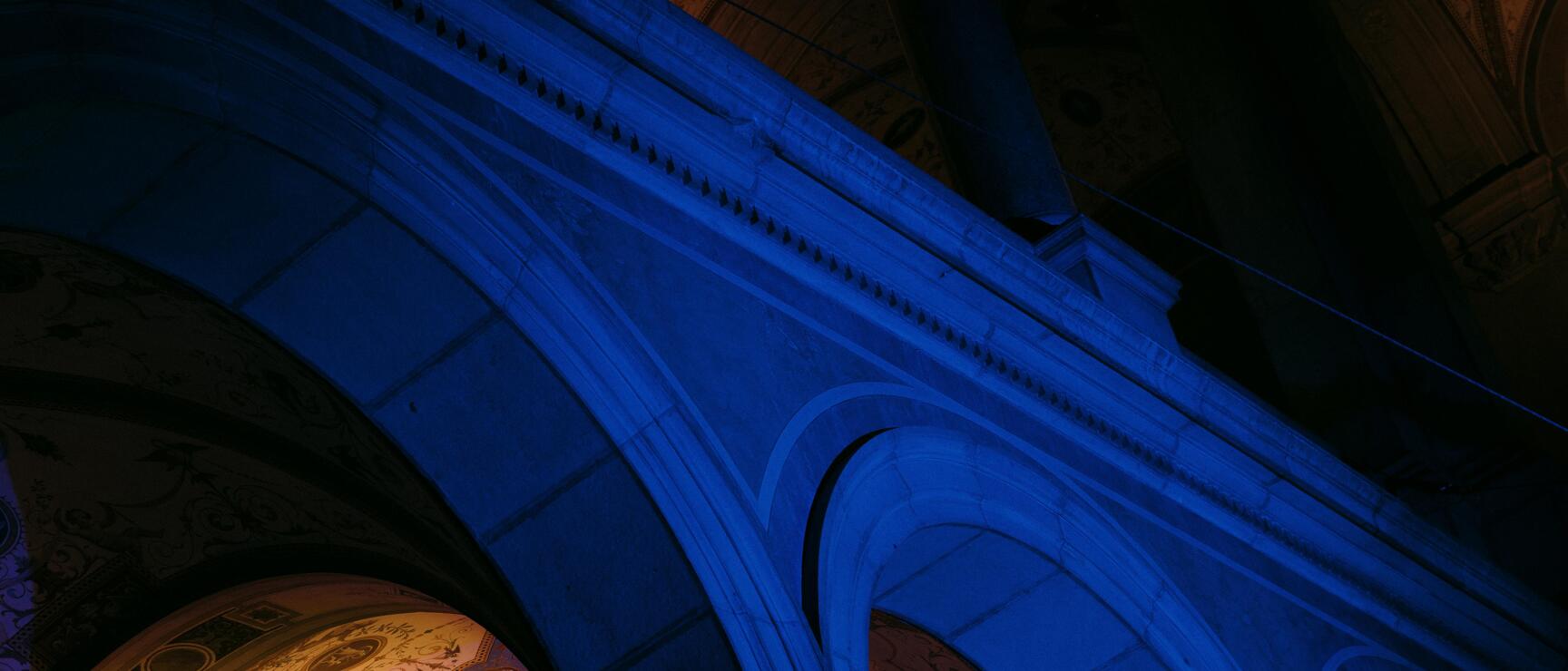 View upward into a barrel vault with frescoes, illuminated in blue and orange, columns and arches visible.