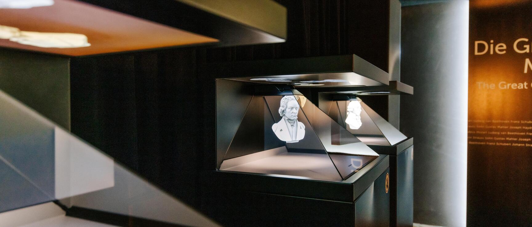 Hologram of a bust in a display case, exhibition room with text "Die Gr..." in background.