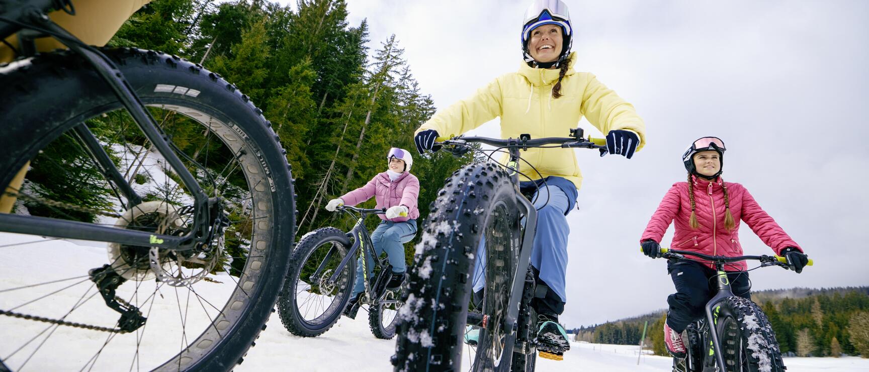 Three women wearing helmets riding fatbikes on a snowy trail with coniferous forest in the background.