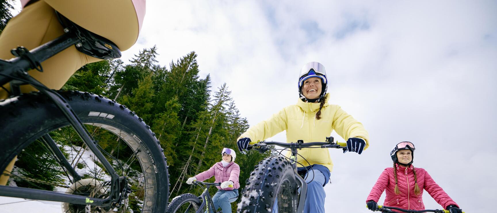 Three women wearing helmets riding fatbikes on a snowy trail with coniferous forest in the background.