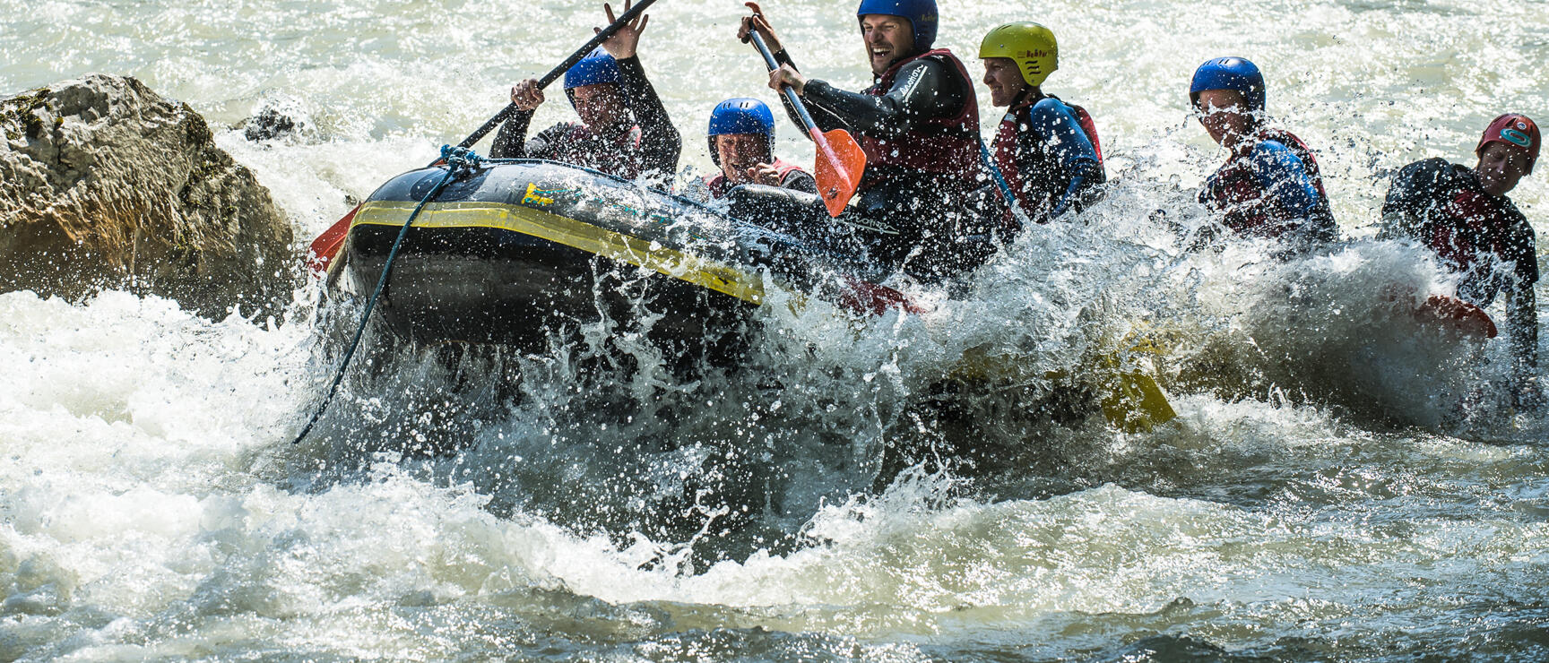 Several rafting boats with people in helmets and wetsuits navigating whitewater rapids with green forest in the background.