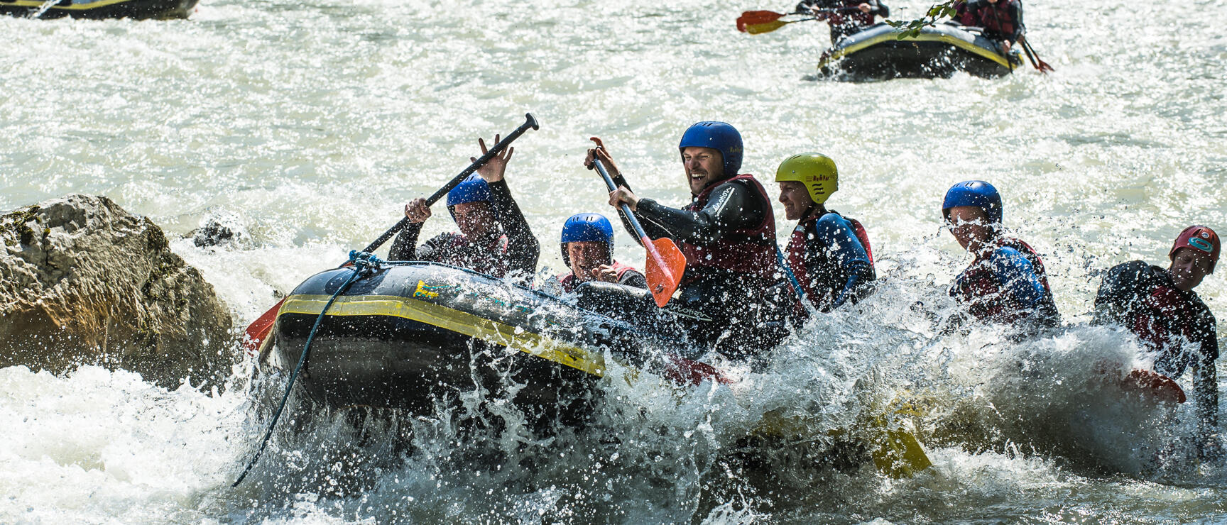 Several rafting boats with people in helmets and wetsuits navigating whitewater rapids with green forest in the background.