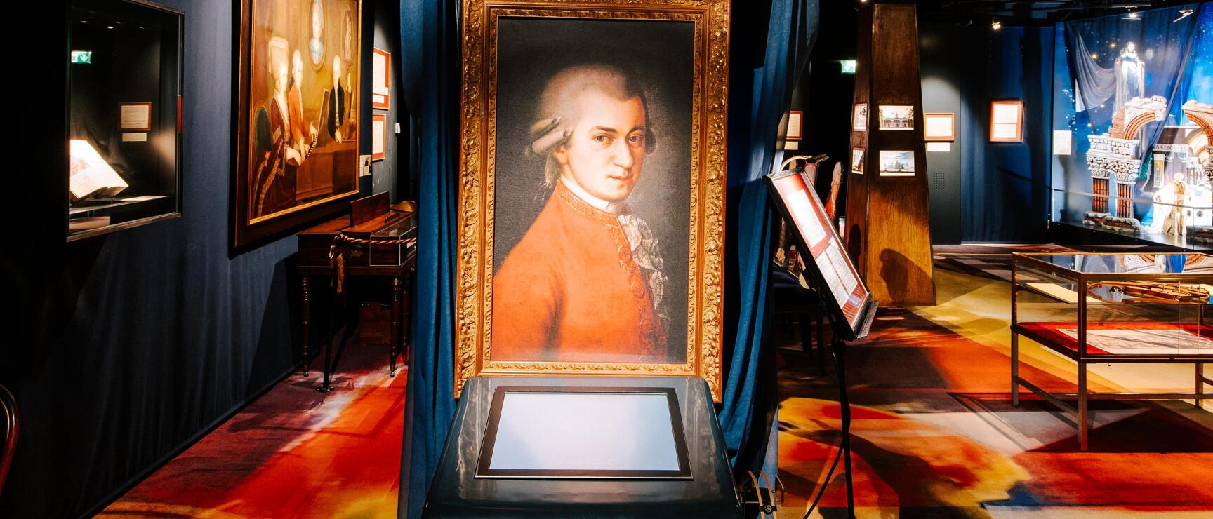 Exhibition room with Mozart portrait in gold frame, 'Amadeus' sign, headphones and colourful carpet.