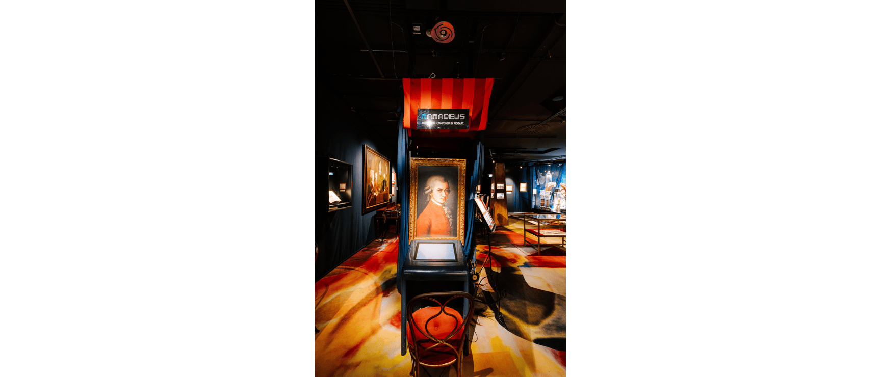 Exhibition room with Mozart portrait in gold frame, 'Amadeus' sign, headphones and colourful carpet.
