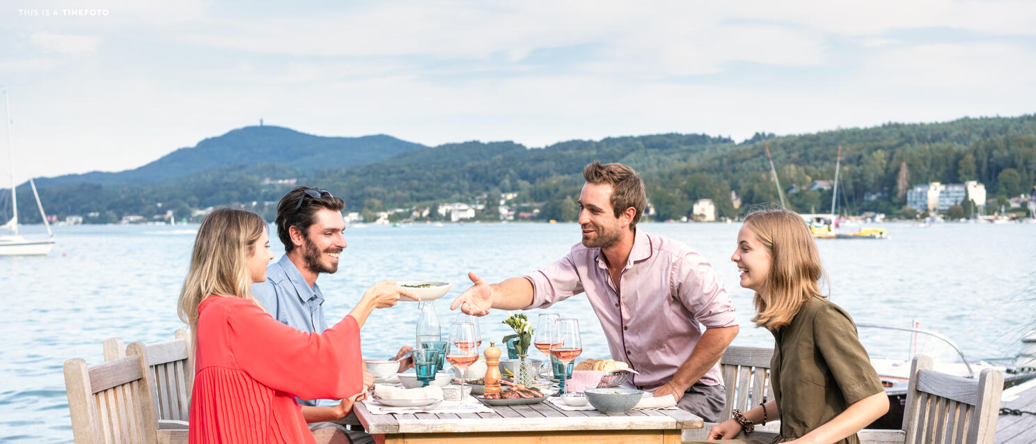 Four people dining at a wooden table directly on Lake Wörthersee with wine and food on the table