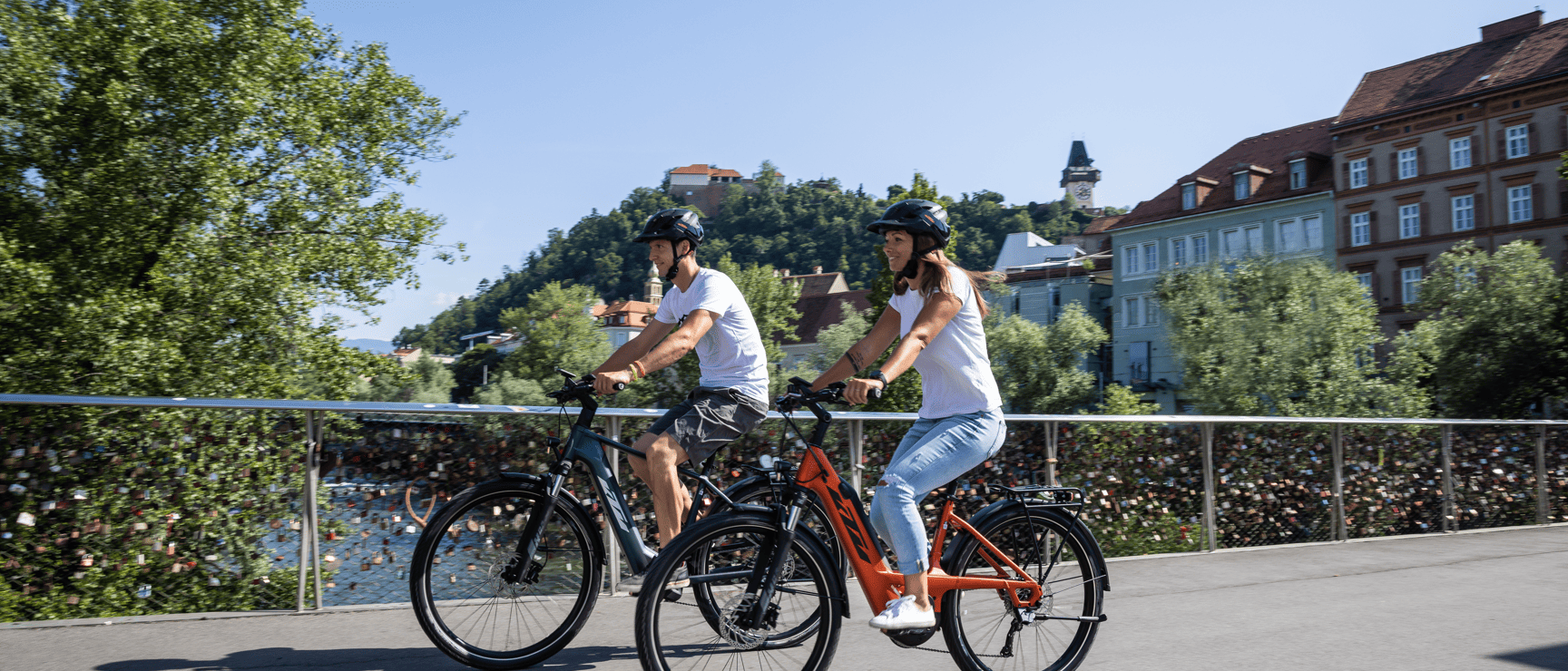 Zwei Radfahrer mit Helmen auf einer Brücke in Graz, Schlossberg mit Uhrturm im Hintergrund.