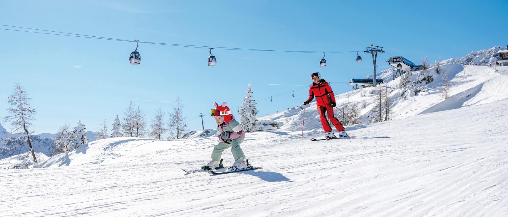 Child and adult in red ski suits on sunny slope with cable car and mountain station in background.