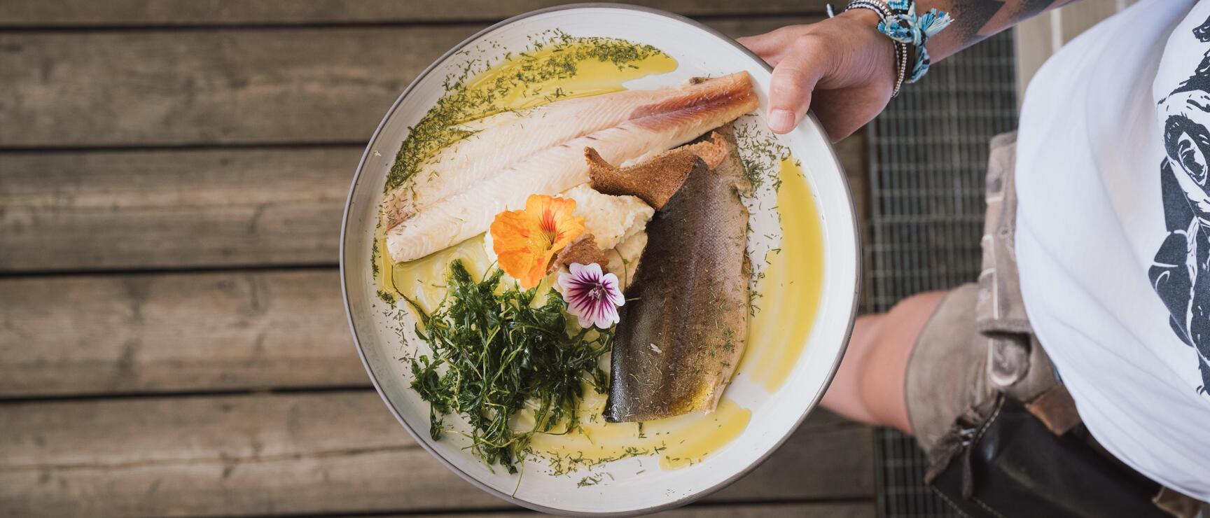 Tattooed hand holding plate with fish fillet, herbs, flower garnish and butter sauce, overhead view.