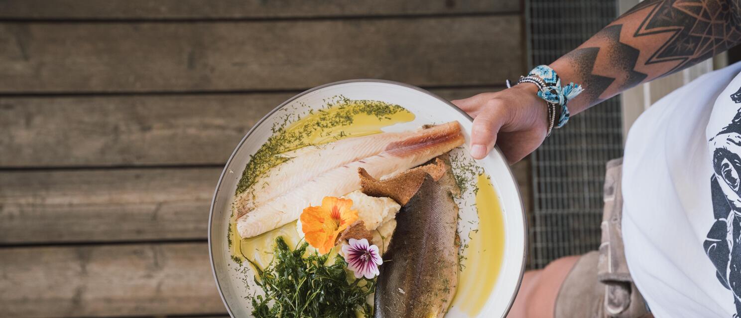 Tattooed hand holding plate with fish fillet, herbs, flower garnish and butter sauce, overhead view.