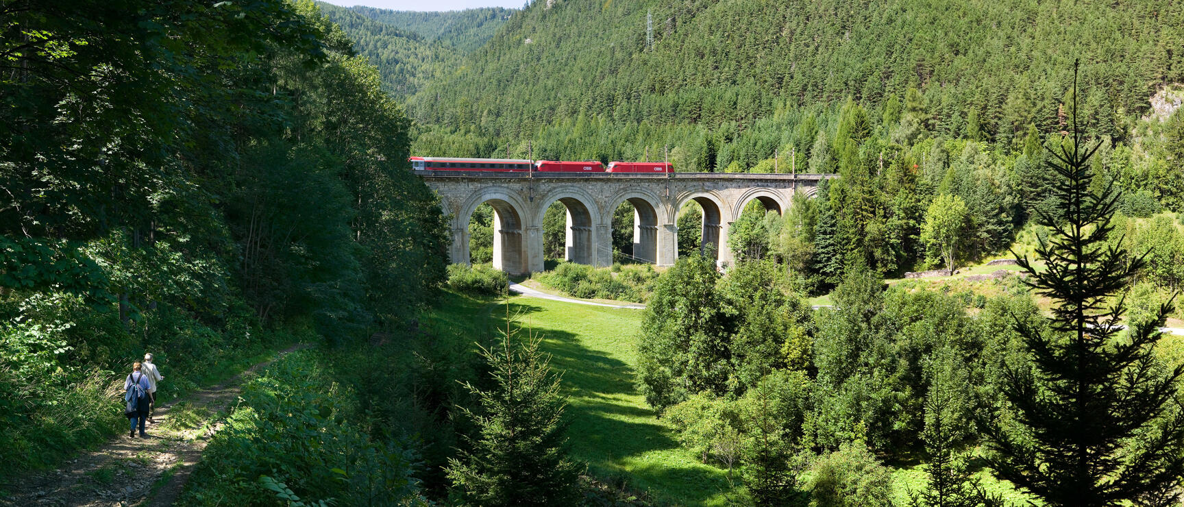 Roter ÖBB-Zug auf dem Semmeringbahn-Viadukt, zwei Wanderer auf Waldweg im Vordergrund.
