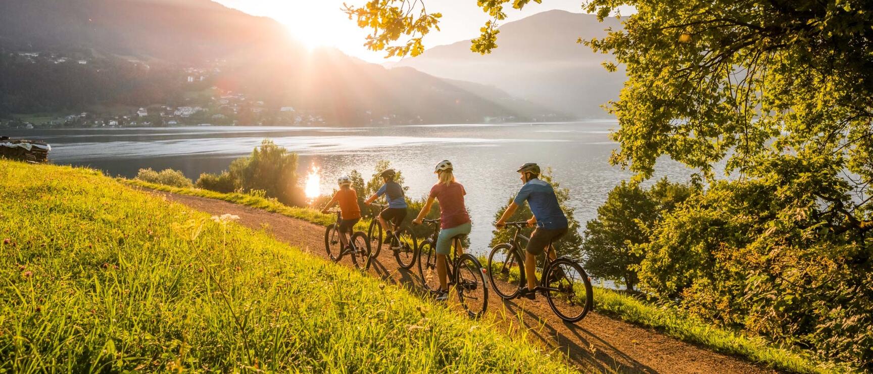 Four cyclists with helmets on a path by a lake, mountains and sunlight in the background.
