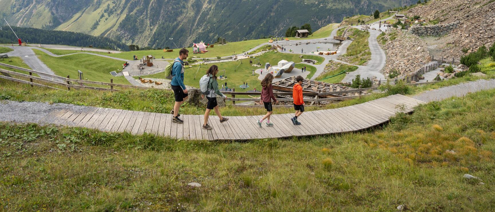 Four people on a wooden jetty in the mountains, cable car and leisure facility in the background.