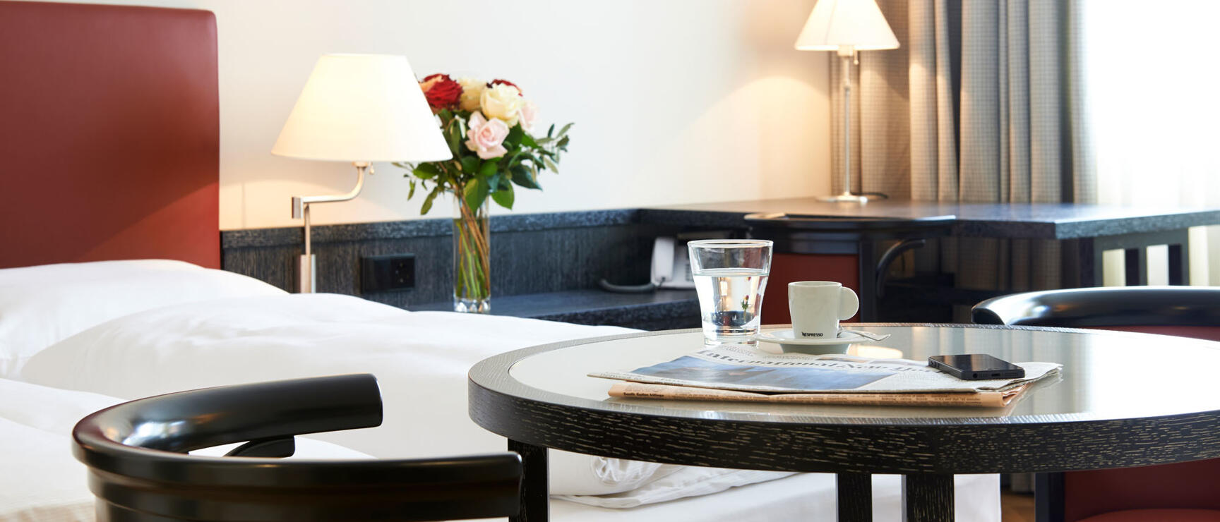 Hotel room with round table, coffee cup, water glass, newspaper, flower bouquet and double bed in background.