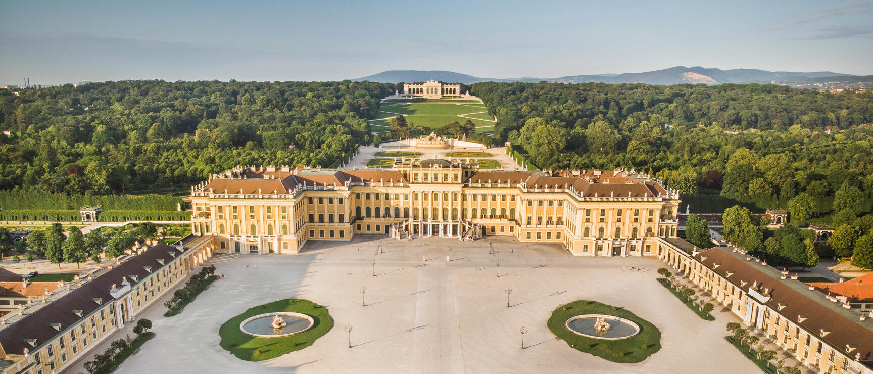 Luftaufnahme von Schloss Schönbrunn in Wien mit Ehrenhof, zwei Brunnen und der Gloriette im Hintergrund.