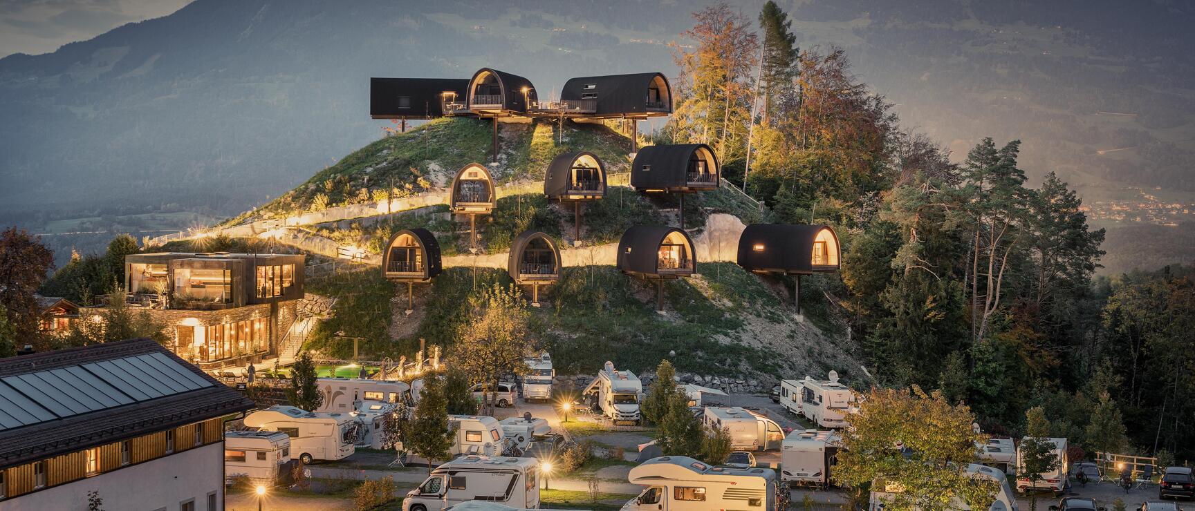 Illuminated glamping pods on a hill at dusk, camping site with caravans below, mountain panorama in background.