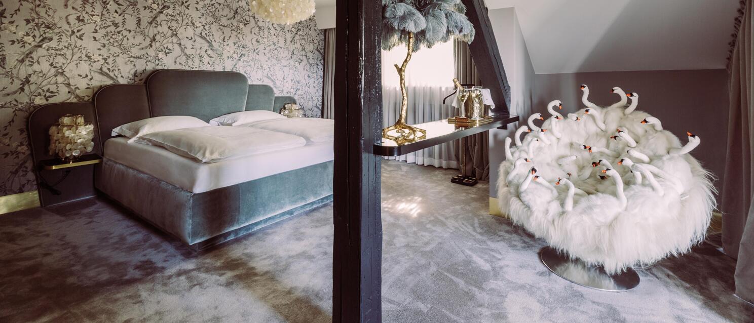 Extravagant hotel room with velvet bed, bird wallpaper, feather lamp and white armchair with swan decoration.