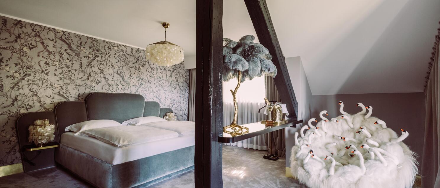 Extravagantes Hotelzimmer mit Samtbett, Vogeltapete, Federlampe und weißem Sessel mit Schwan-Dekor.